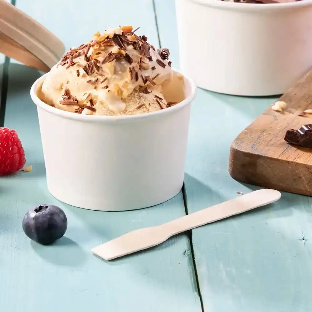 White disposable spoon next to a cup of ice cream topped with chocolate and nuts, with berries on a blue wooden surface.