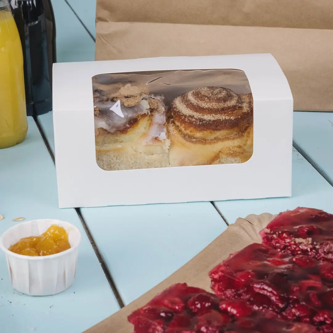White cake box with a clear window containing two cinnamon rolls, next to a cup of yellow jam and a slice of red dessert.