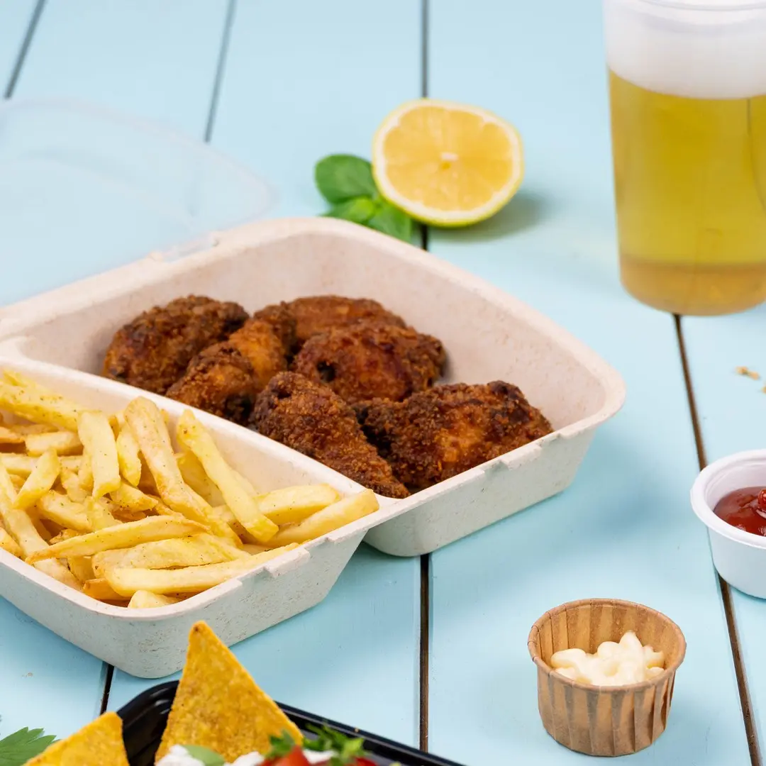 Takeout container with fried chicken and fries, lemon slice, beer, and dipping sauces on a blue wooden surface.