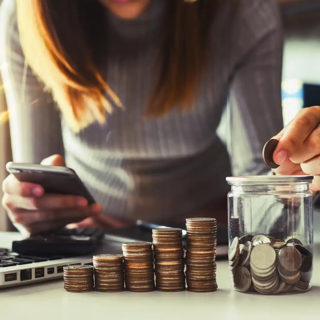 Woman placing a coin into a glass jar while using a smartphone, with stacks of coins on a table.