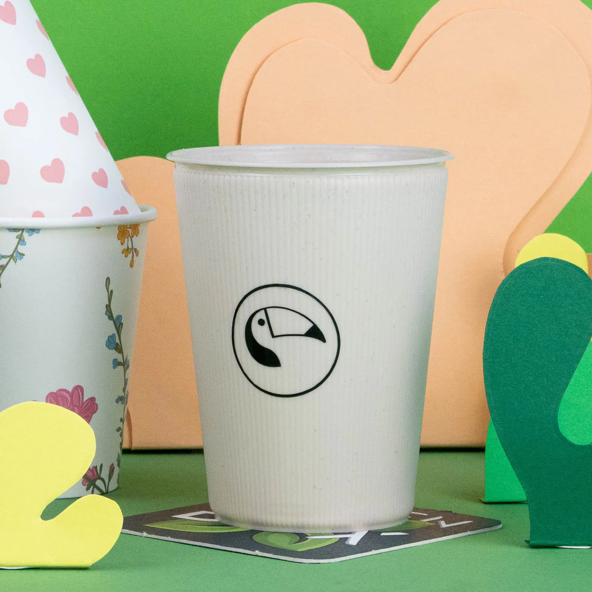White ripple reusable cup with a toucan logo, placed among colorful paper decorations on a green background.