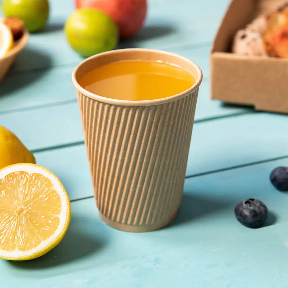 A ribbed cardboard cup filled with yellow liquid, surrounded by lemon slices, fruits, and a box of baked goods.