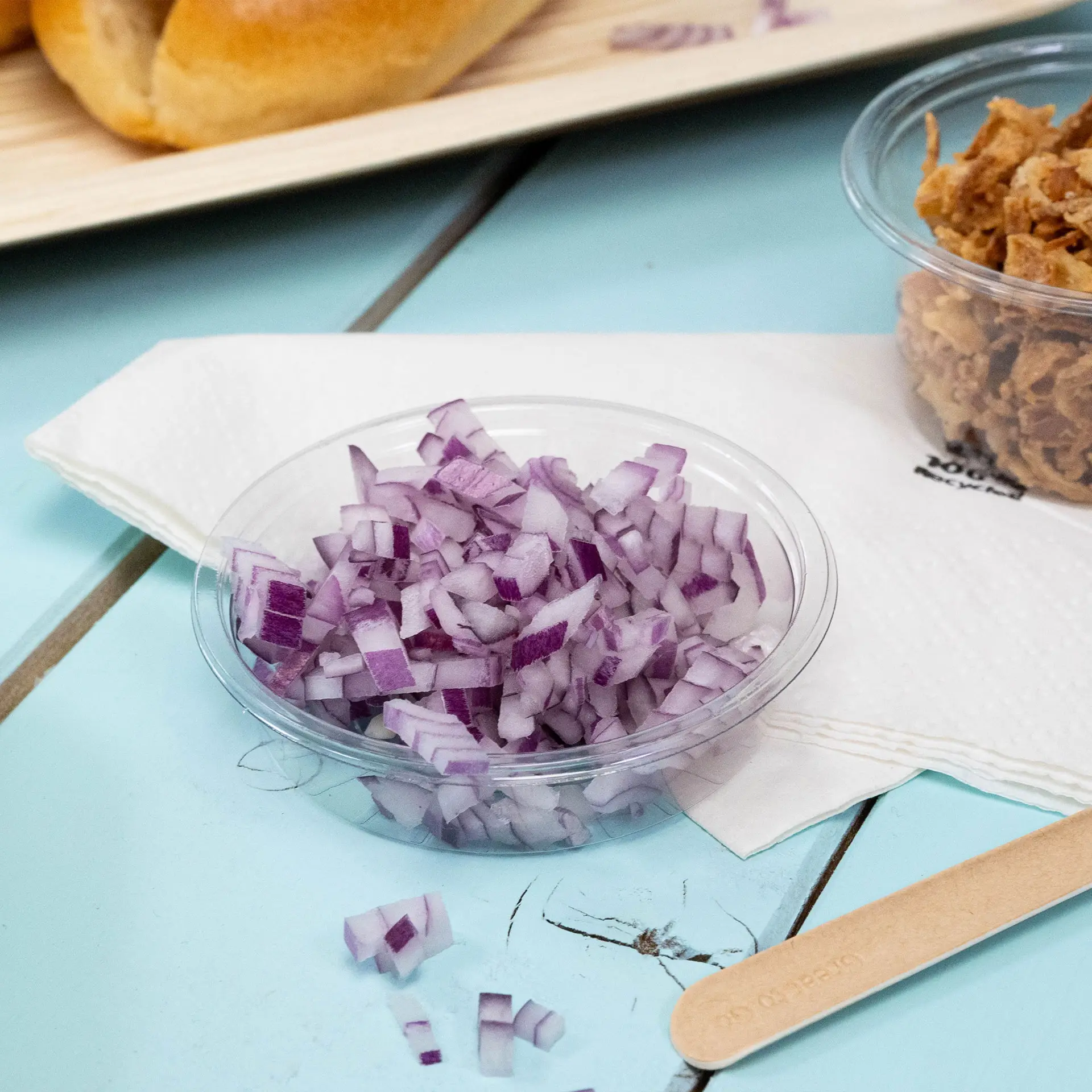 Chopped red onions in a clear portion cup on a blue surface, with a napkin and a wooden stir stick nearby.