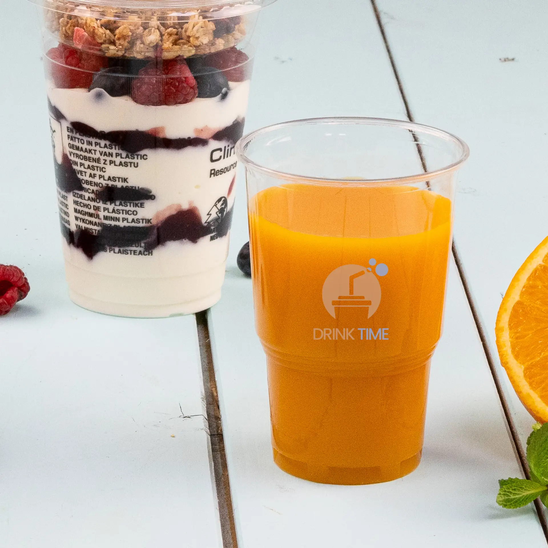 One clear rPET cup filled with orange juice, featuring a blue diamond logo; served with fresh orange and mint leaves nearby.