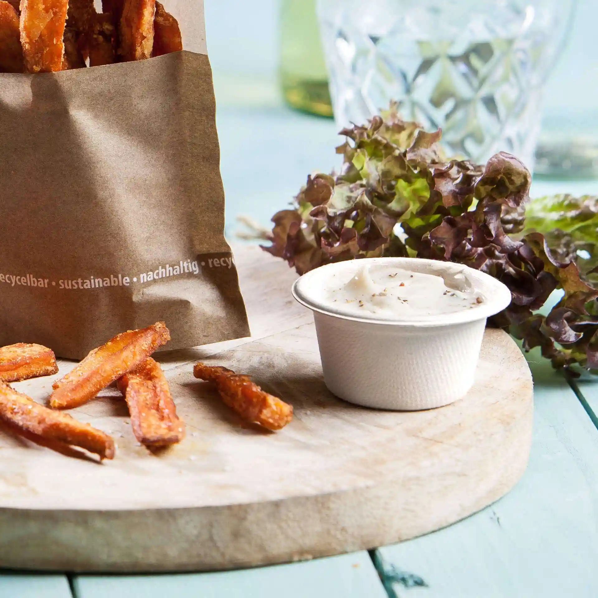 Image of a paper bag with sweet potato fries, a white dip bowl, and green lettuce on a wooden board.