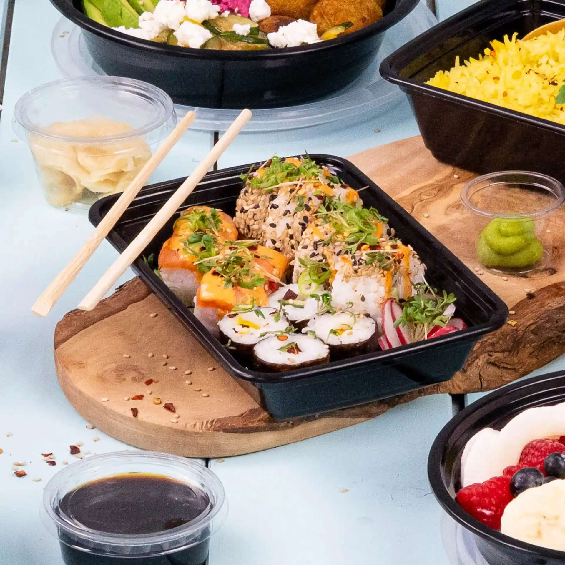 Sushi rolls with toppings in a black rectangular reusable container, accompanied by chopsticks and condiments.