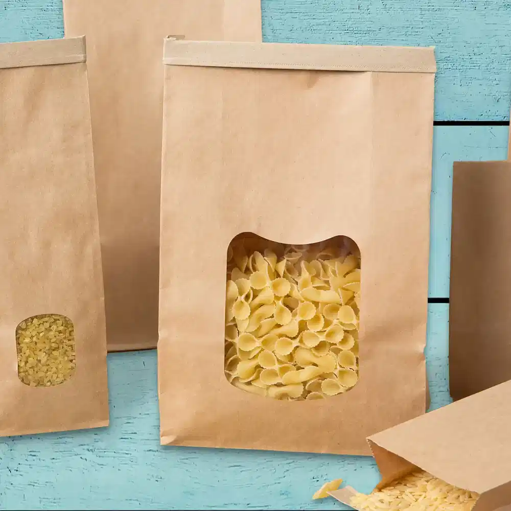 Brown paper bags with transparent windows, filled with various pasta shapes, on a blue wooden surface.