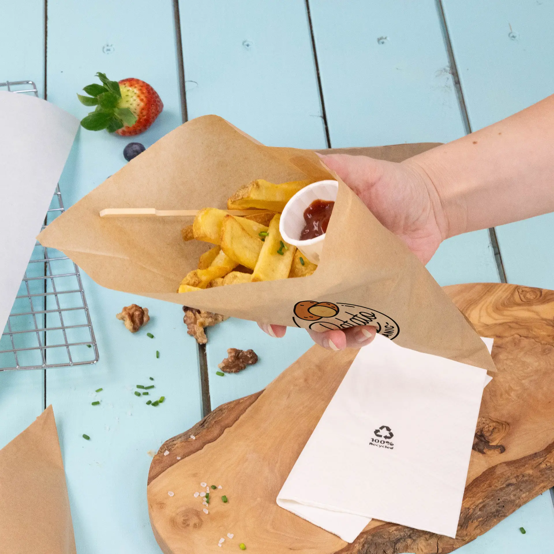 Hand holding a brown paper cone filled with French fries and a small cup of ketchup, placed on a blue wooden surface.