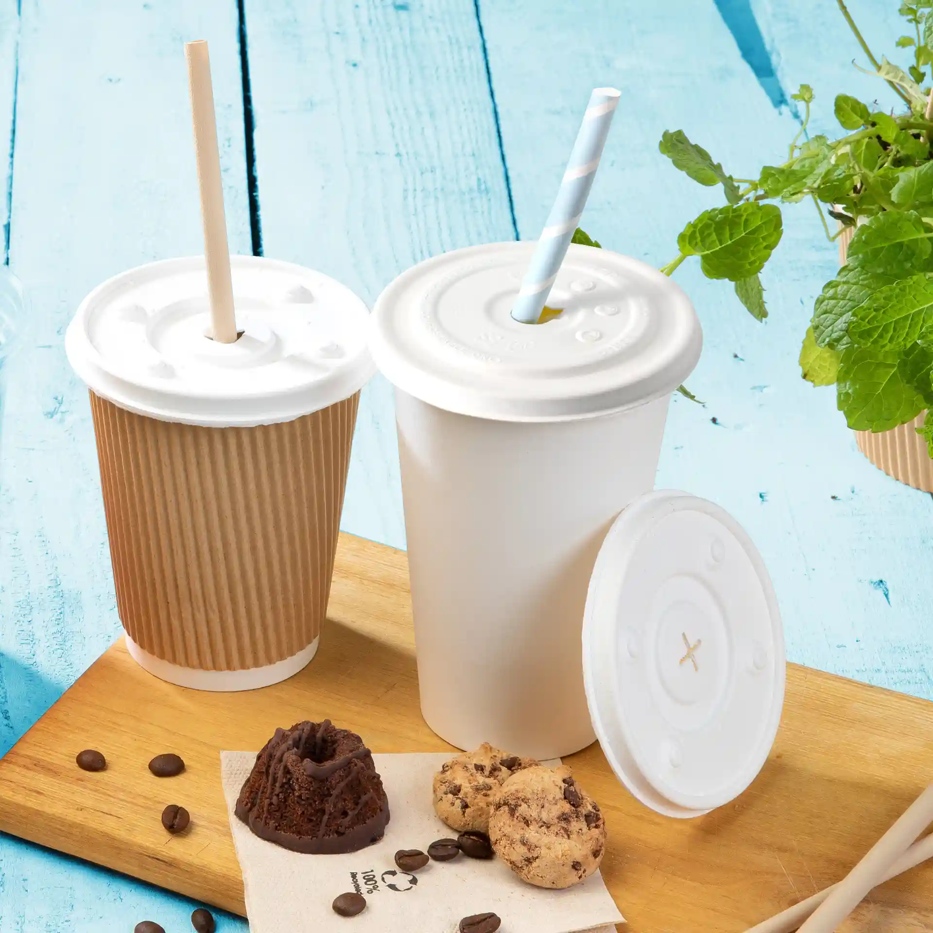 Image of two cups with white lids, one with a straw, placed on a wooden surface with cookies and coffee beans.