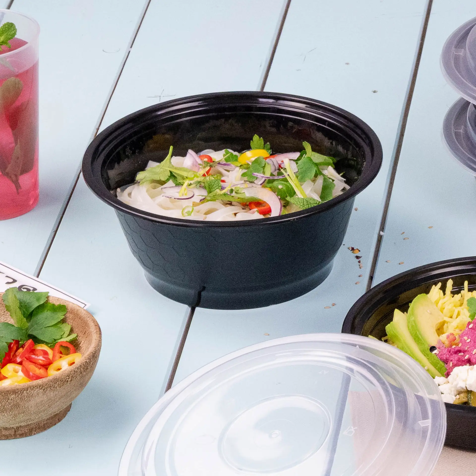 A black reusable food container filled with noodles and garnished with herbs and chilies, surrounded by other food items.