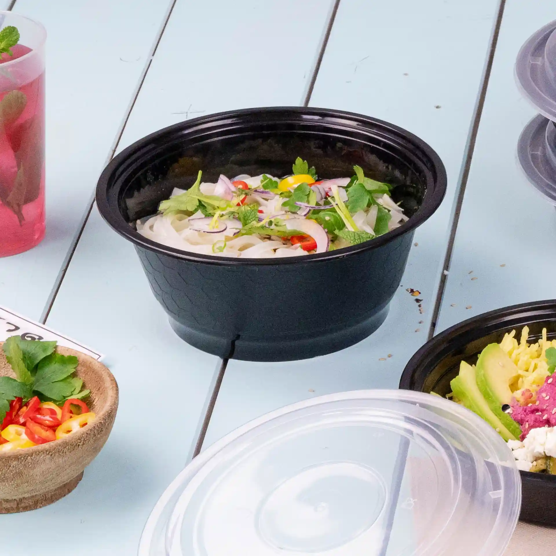 Black reusable container filled with noodles, garnished with herbs and vegetables, on a light blue surface.