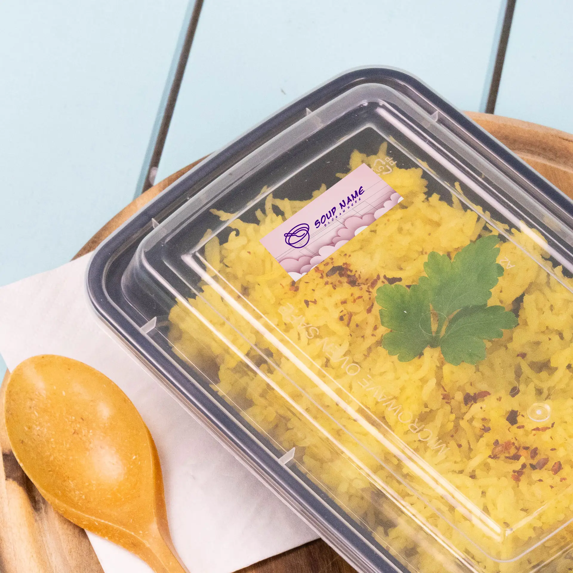 A clear food container with yellow rice and a green herb garnish, covered with a printed label. A wooden spoon is beside it.
