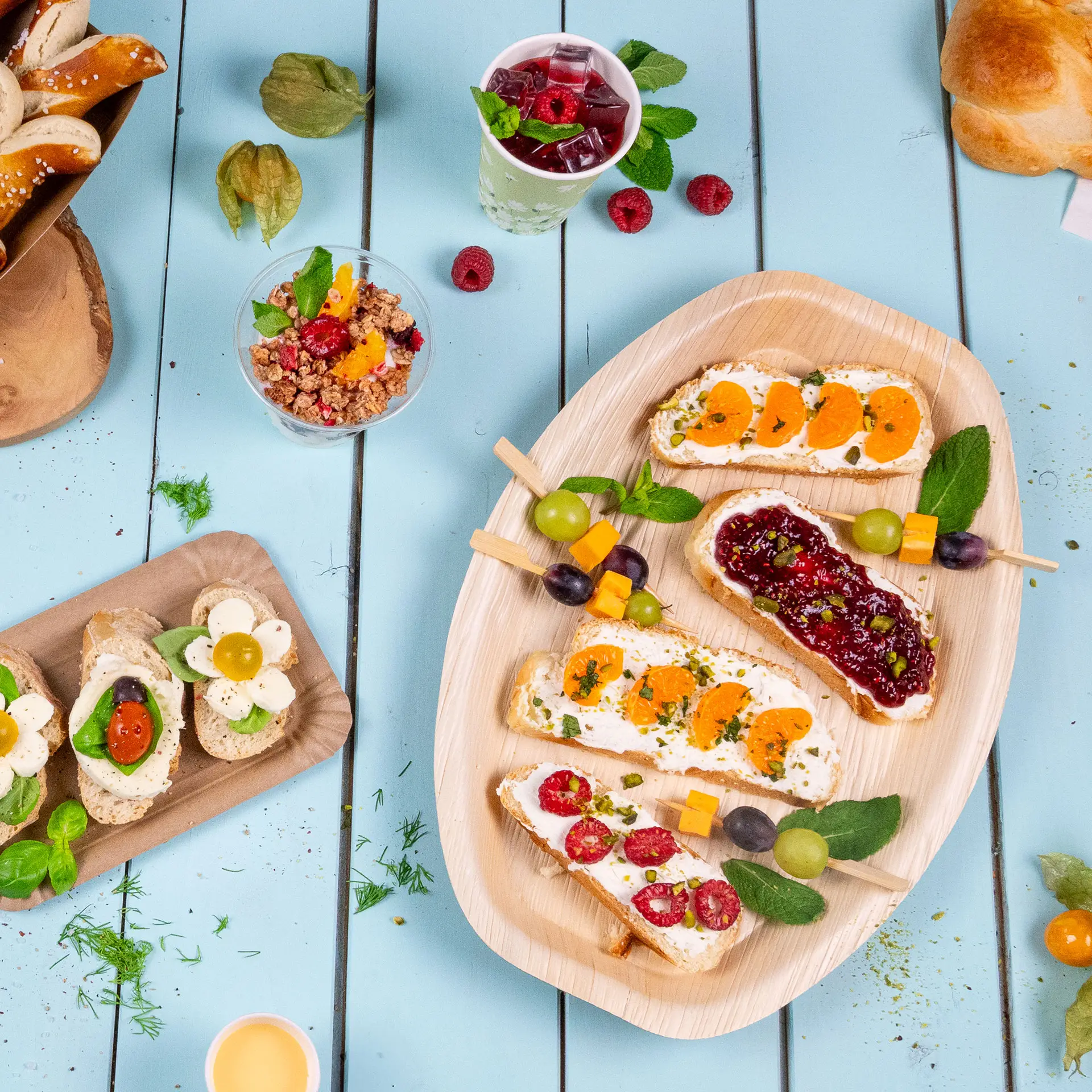 One palm leaf tray with various finger foods including cream cheese topped with fruits, arranged elegantly; fruit and beverage items nearby.
