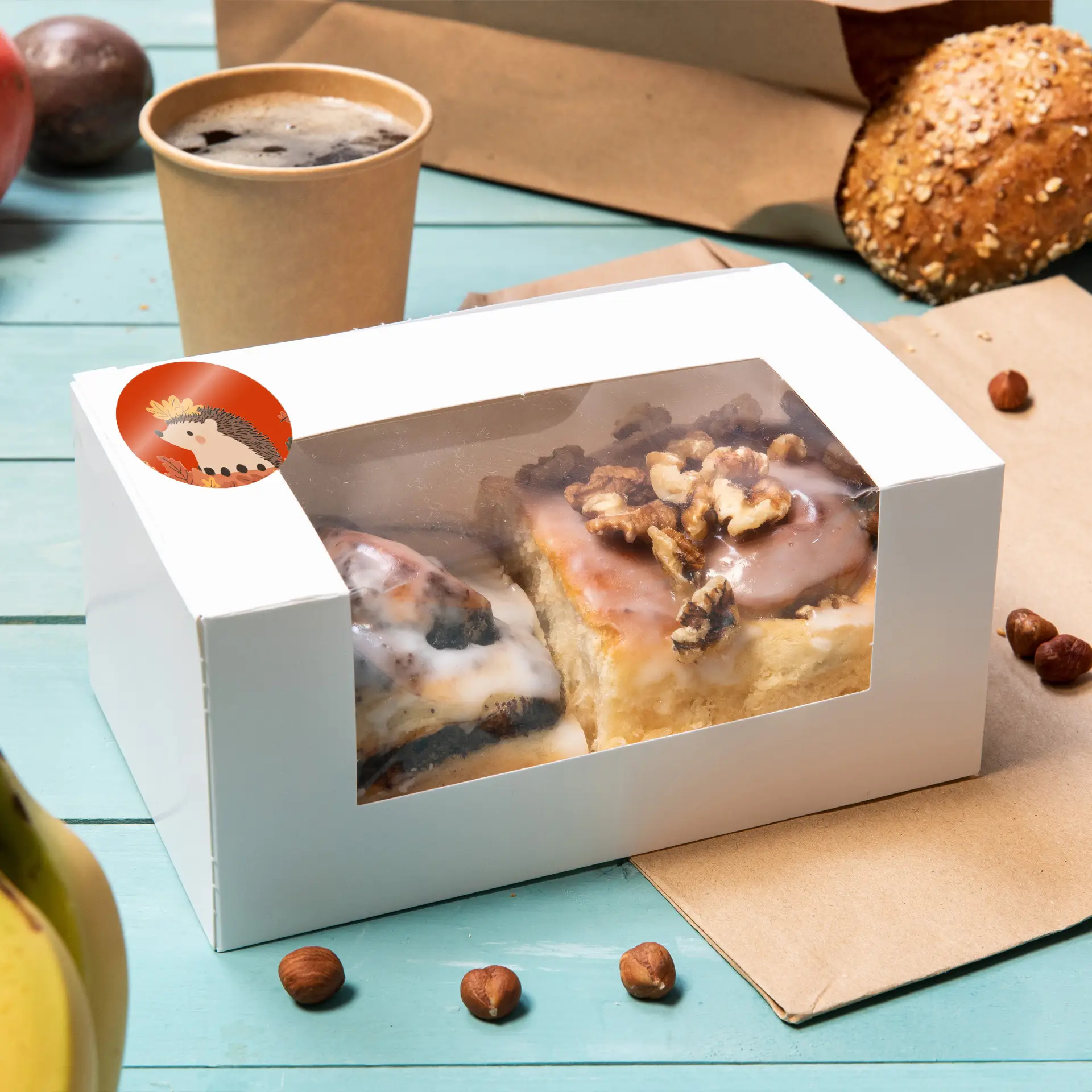 White box with a clear lid containing two baked goods, topped with icing and nuts, on a blue wooden surface.