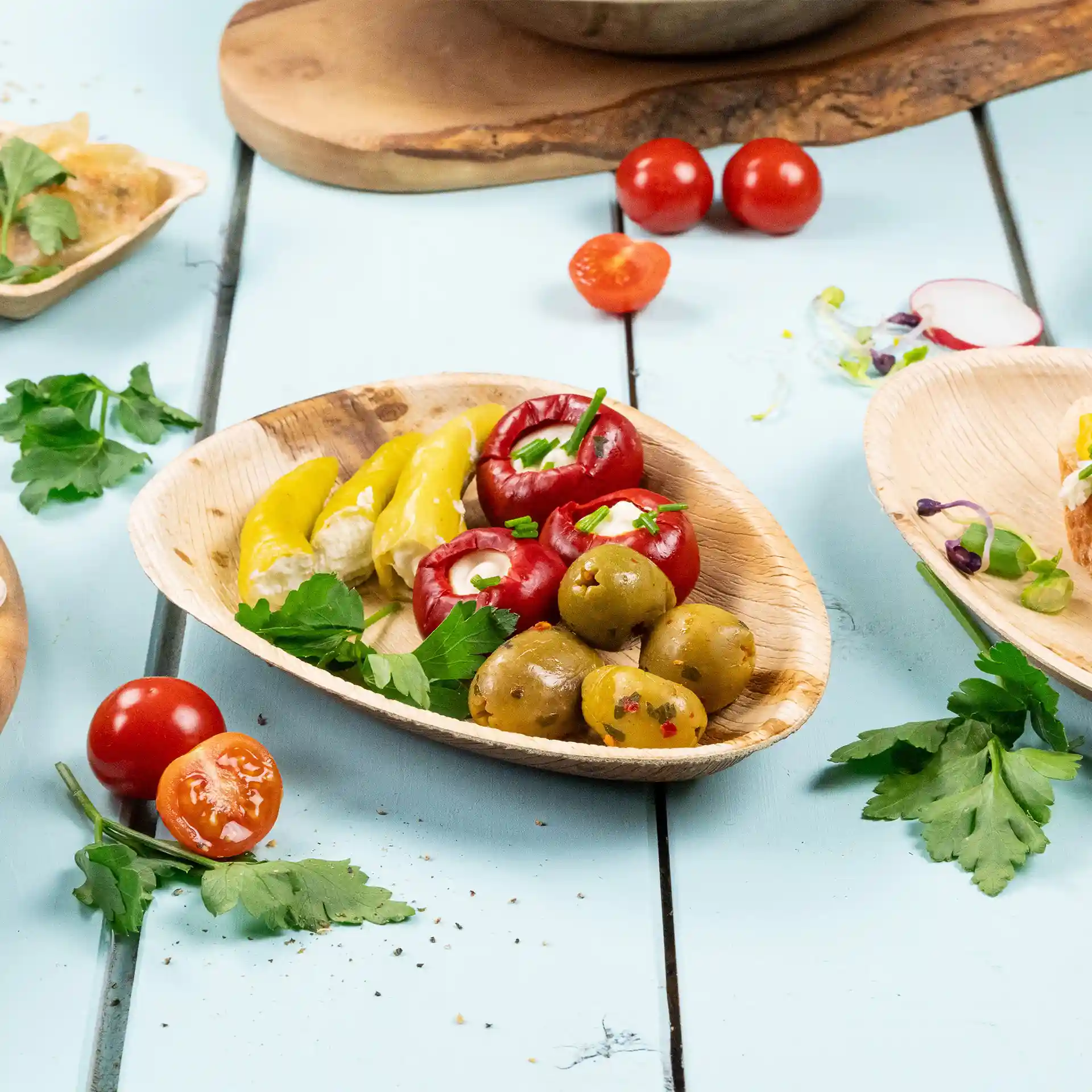 Oval palm leaf plate with yellow peppers, red stuffed tomatoes, green olives, and parsley on a blue surface.