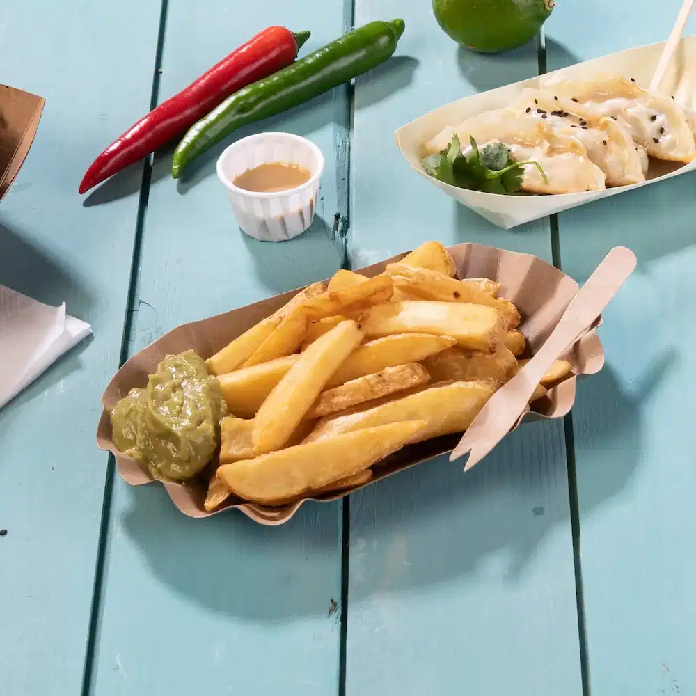 Basket of French fries with green sauce, wooden utensils, and chili peppers on a blue wooden table.