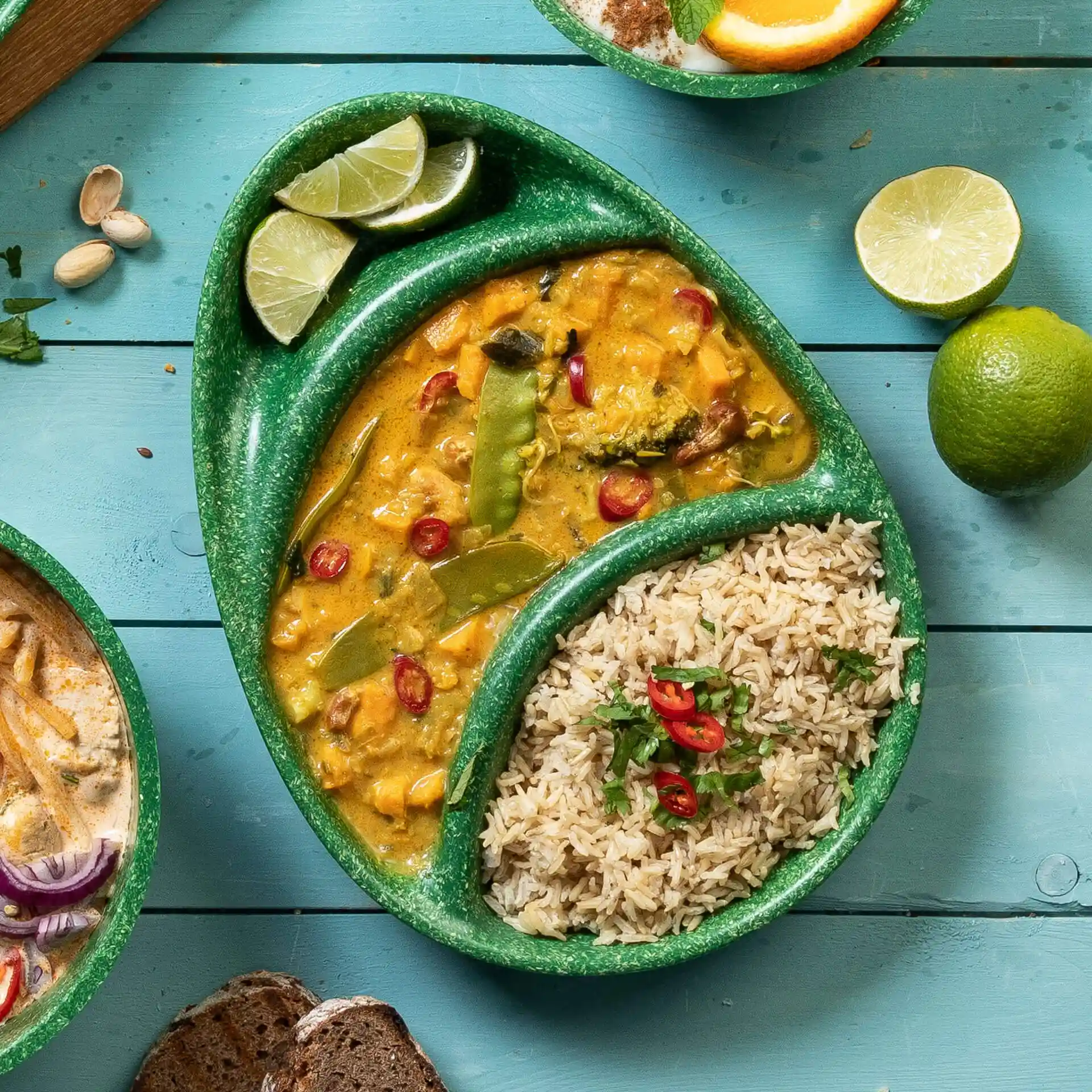 Green reusable plate with three compartments, filled with curry, rice, and lime wedges on a blue wooden surface.