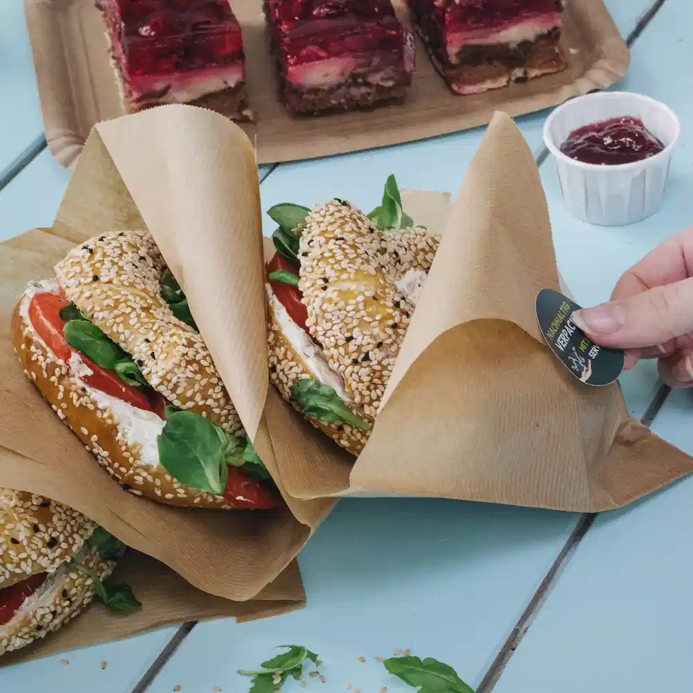 Two sesame-seed buns filled with lettuce and tomato, wrapped in paper; a hand holds a label, with dessert in the background.