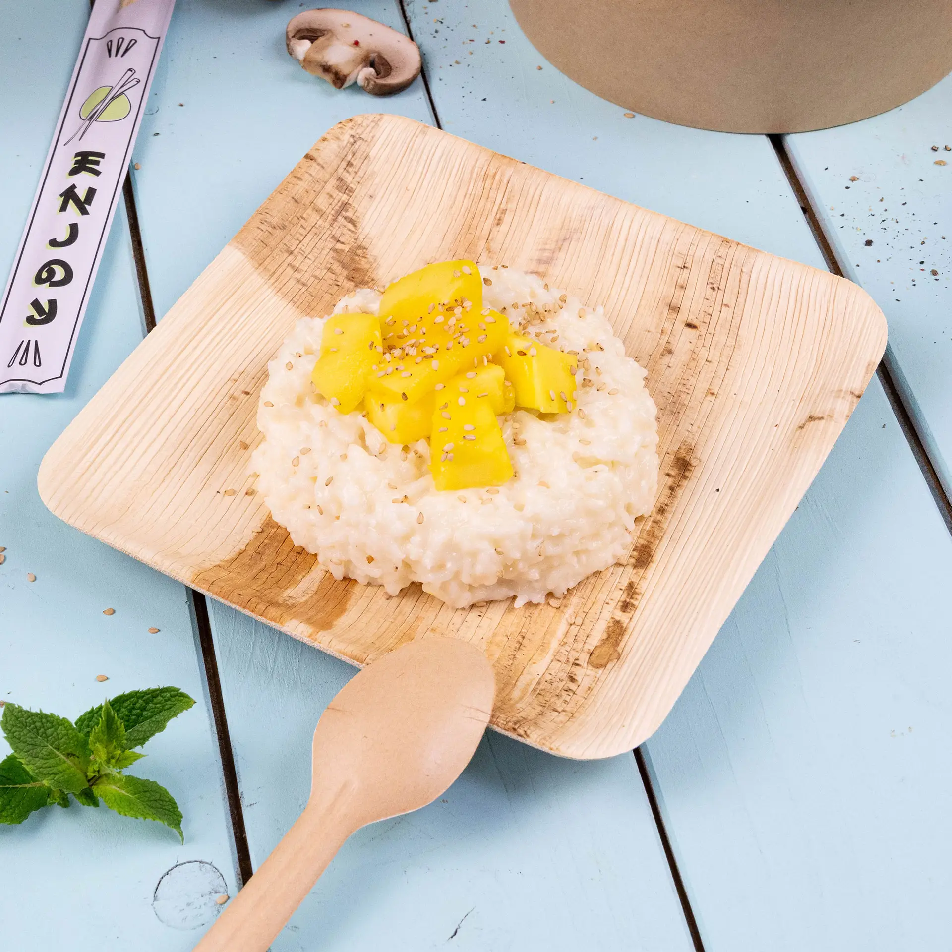 Square palm leaf plate with risotto topped with mango pieces and sesame seeds, on a blue background with a wooden spoon.