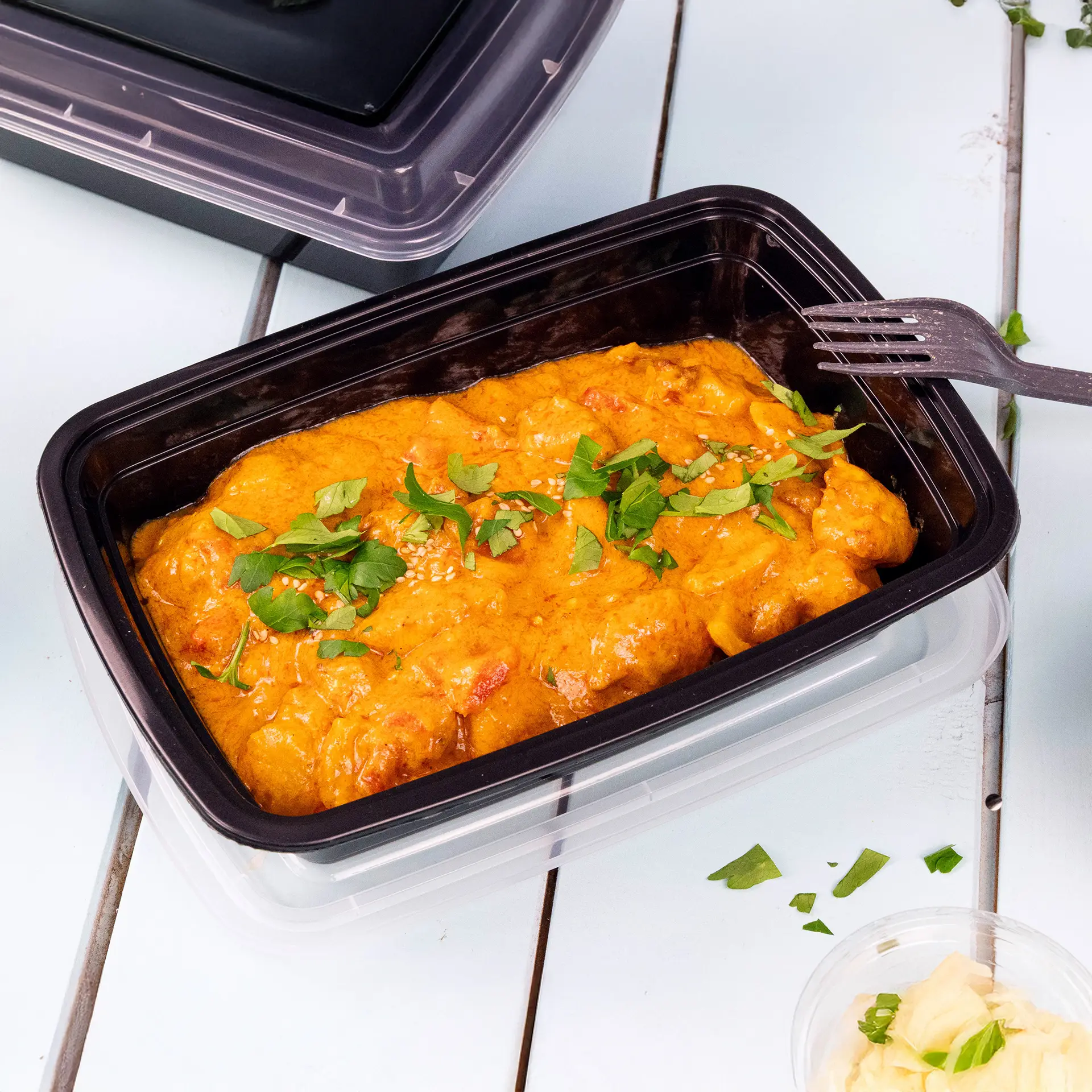 Reusable black PP container with 1000 ml capacity, filled with a curry dish and garnished with herbs; fork beside the container.