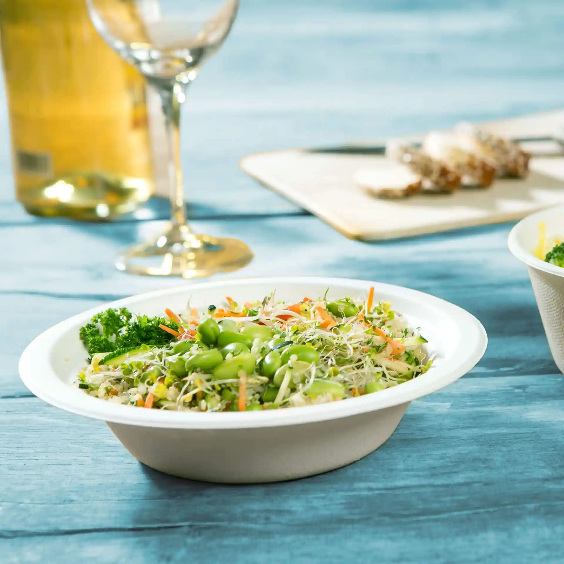 Round white sugarcane bowl filled with mixed salad, including edamame, carrots, and sprouts, on a blue wooden surface.