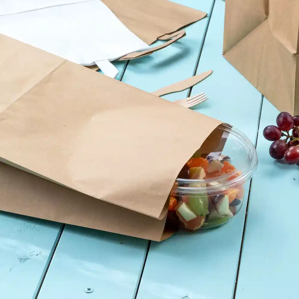 Brown paper bags partially covering a clear container of salad on a blue wooden table. Grapes are visible nearby.