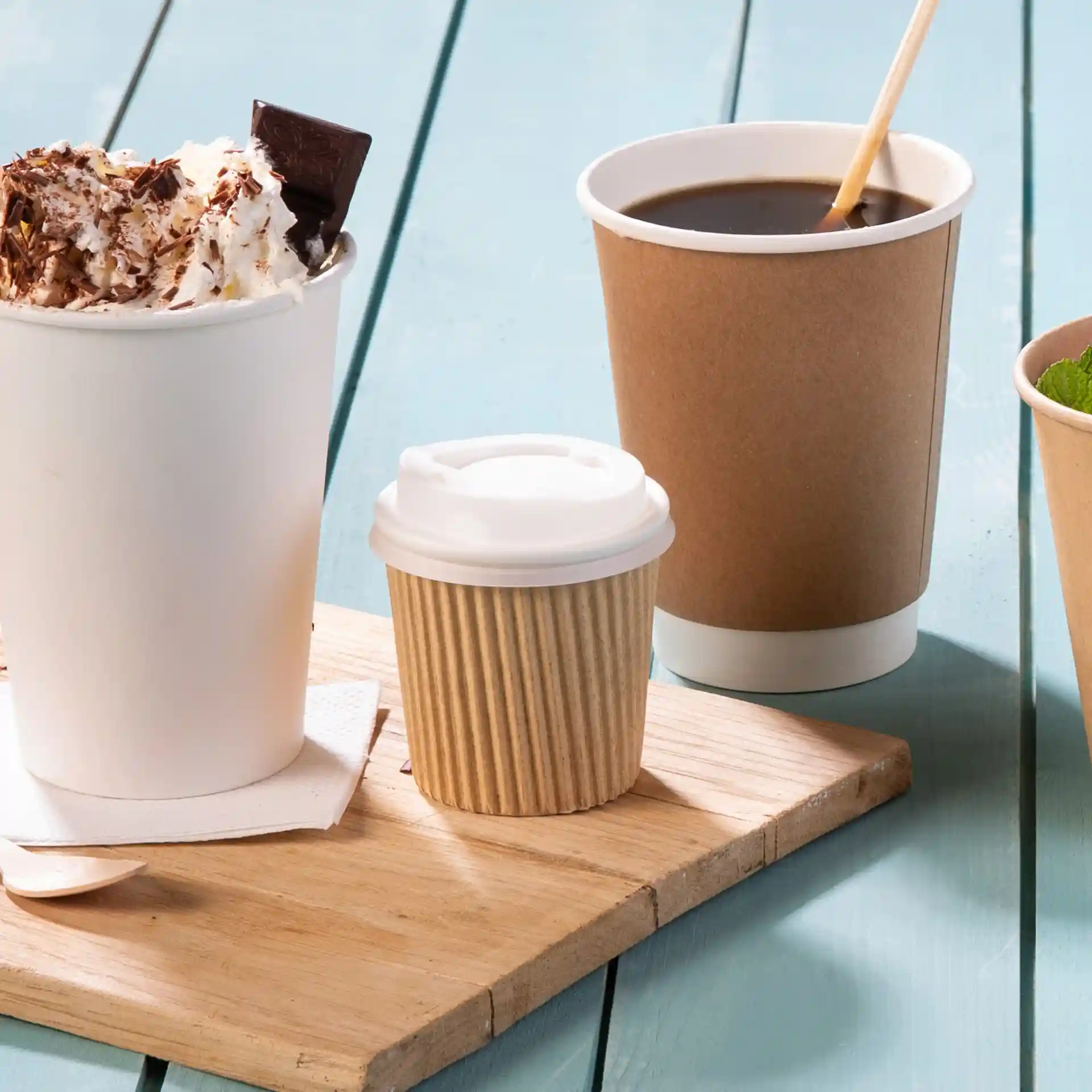 Image of a wooden board with a large white cup topped with whipped cream and chocolate, a small ribbed brown cup, and a brown coffee cup.