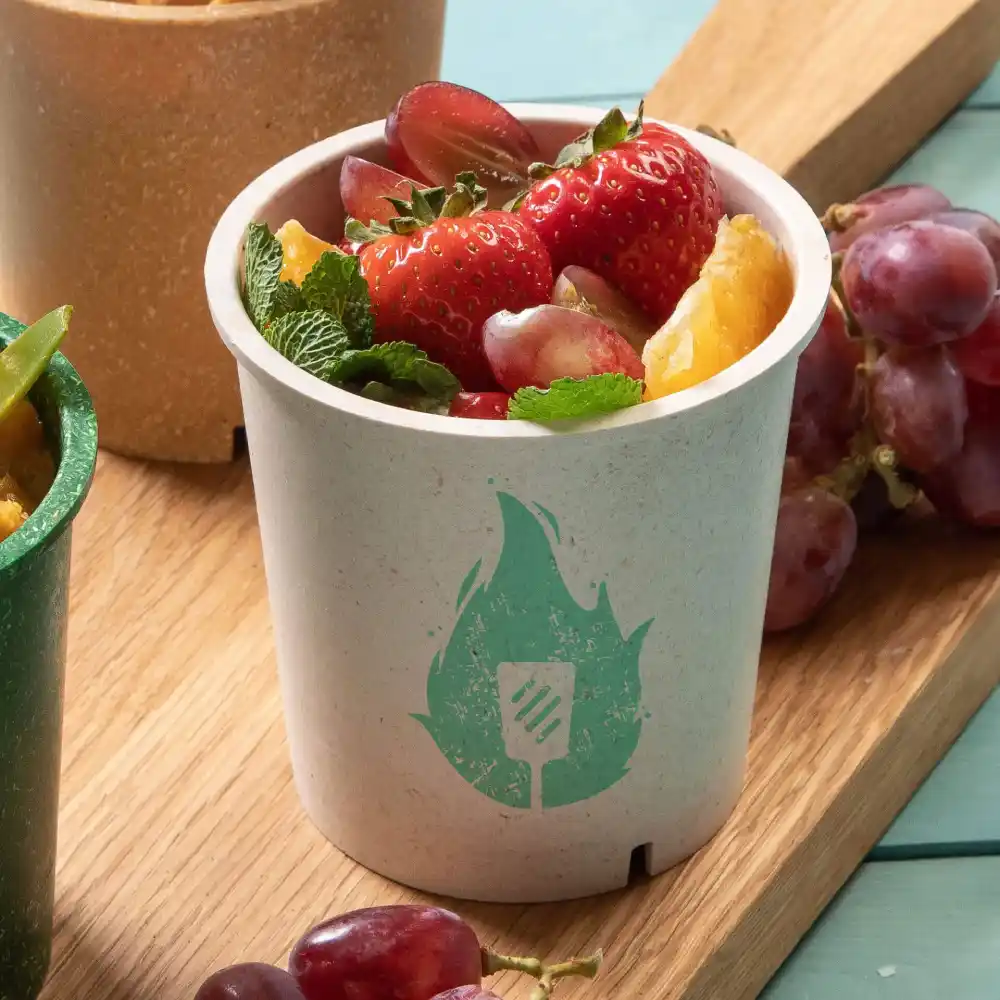 White cup filled with mixed fruits, featuring a green flame logo, next to a green cup and grapes on a wooden surface.