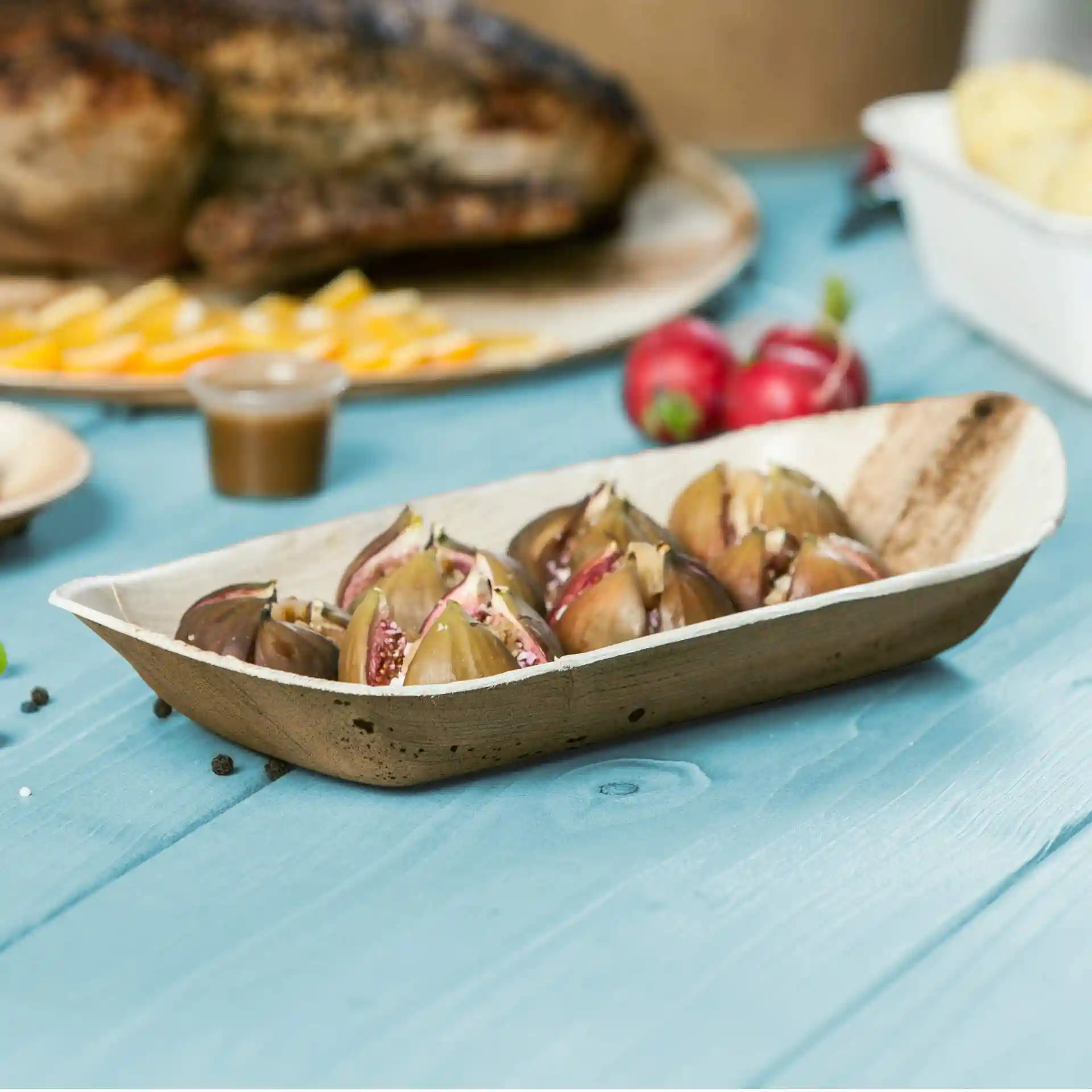Deep palm leaf boat filled with sliced figs, placed on a blue wooden surface with other food items in the background.
