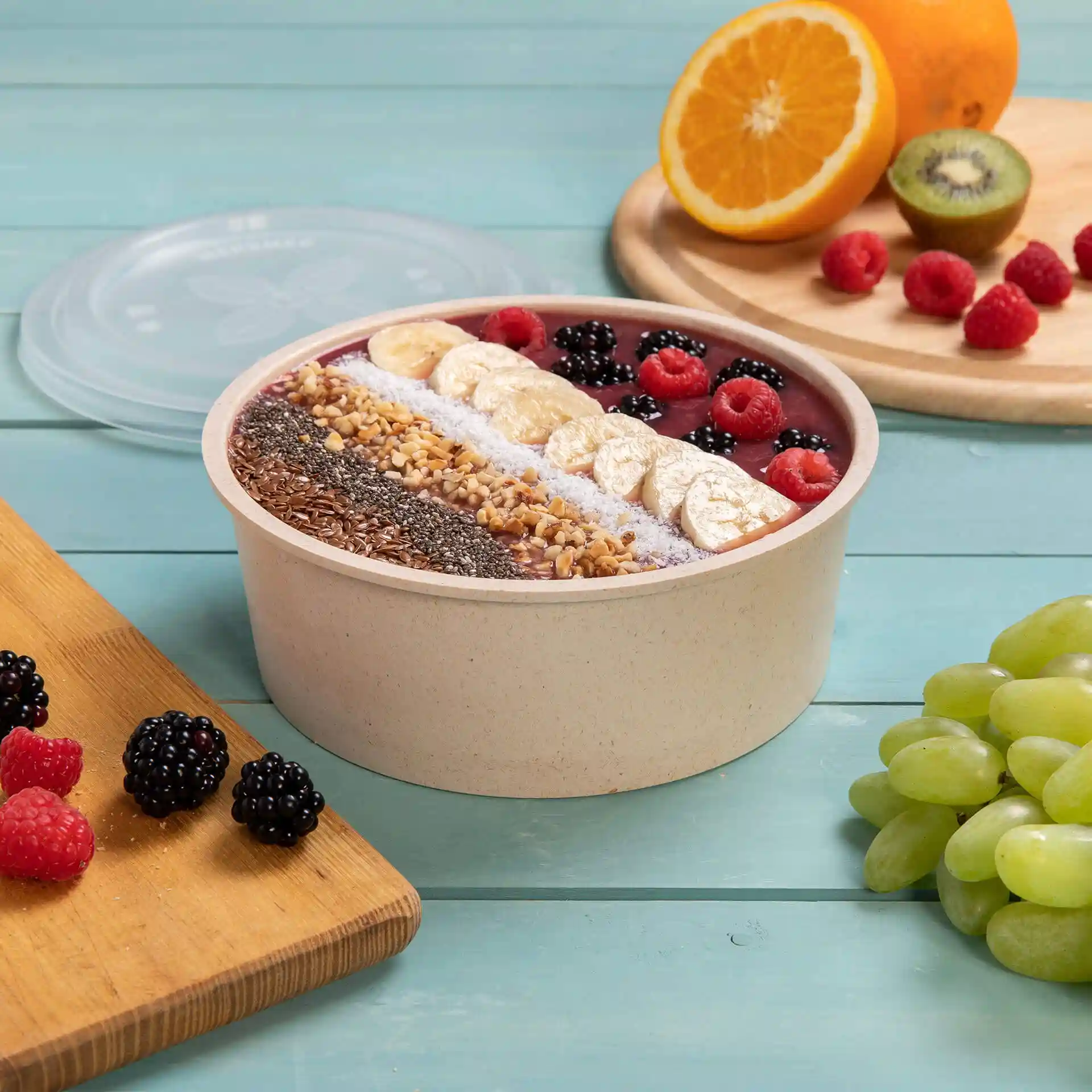 Round reusable bowl filled with smoothie and topped with fruits, placed on a blue wooden surface with additional fruits nearby.
