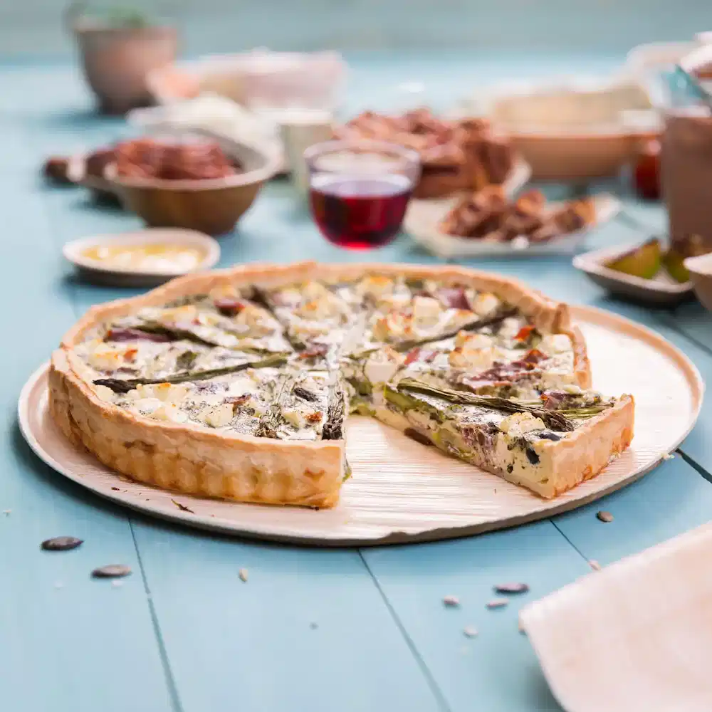 Round vegetable tart on a wooden plate, with a slice removed, set on a turquoise table with various dishes in the background.