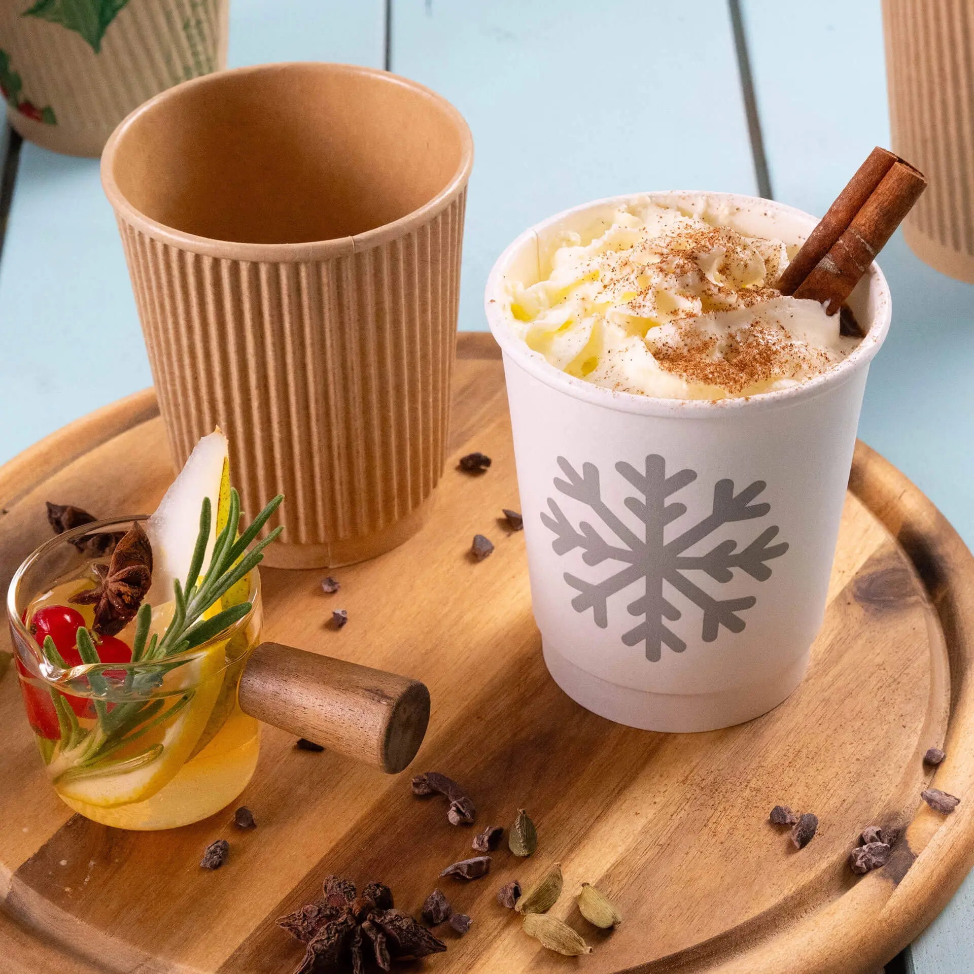 One white insulated coffee cup with a snowflake design filled with whipped cream and a cinnamon stick, next to a ribbed brown cup and a small glass with a garnish.