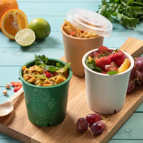 Three reusable soup cups on a wooden board, filled with soup, fruit, and noodles, surrounded by fresh citrus and grapes.