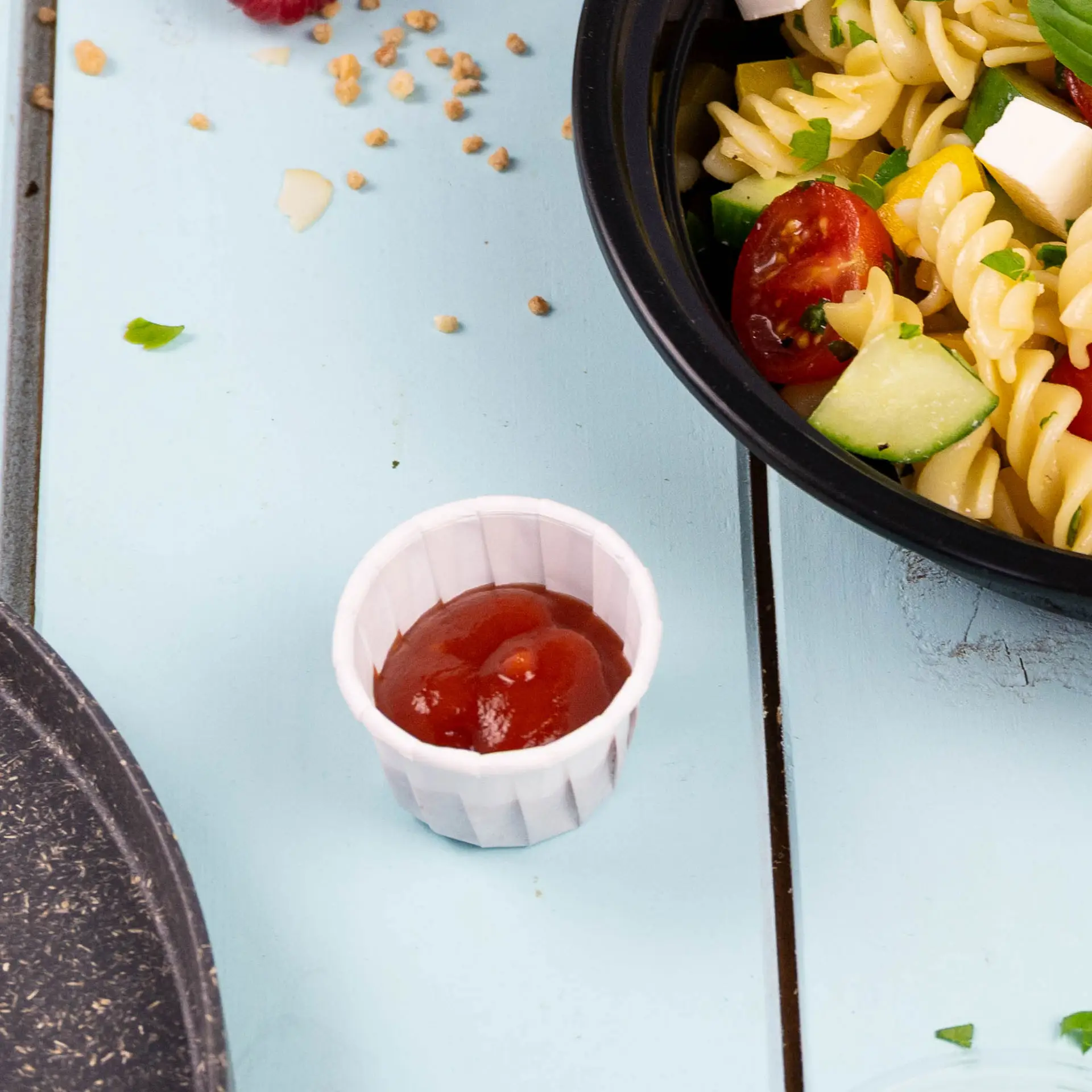 Small white paper cup filled with ketchup, placed beside a bowl of pasta salad on a blue wooden surface.