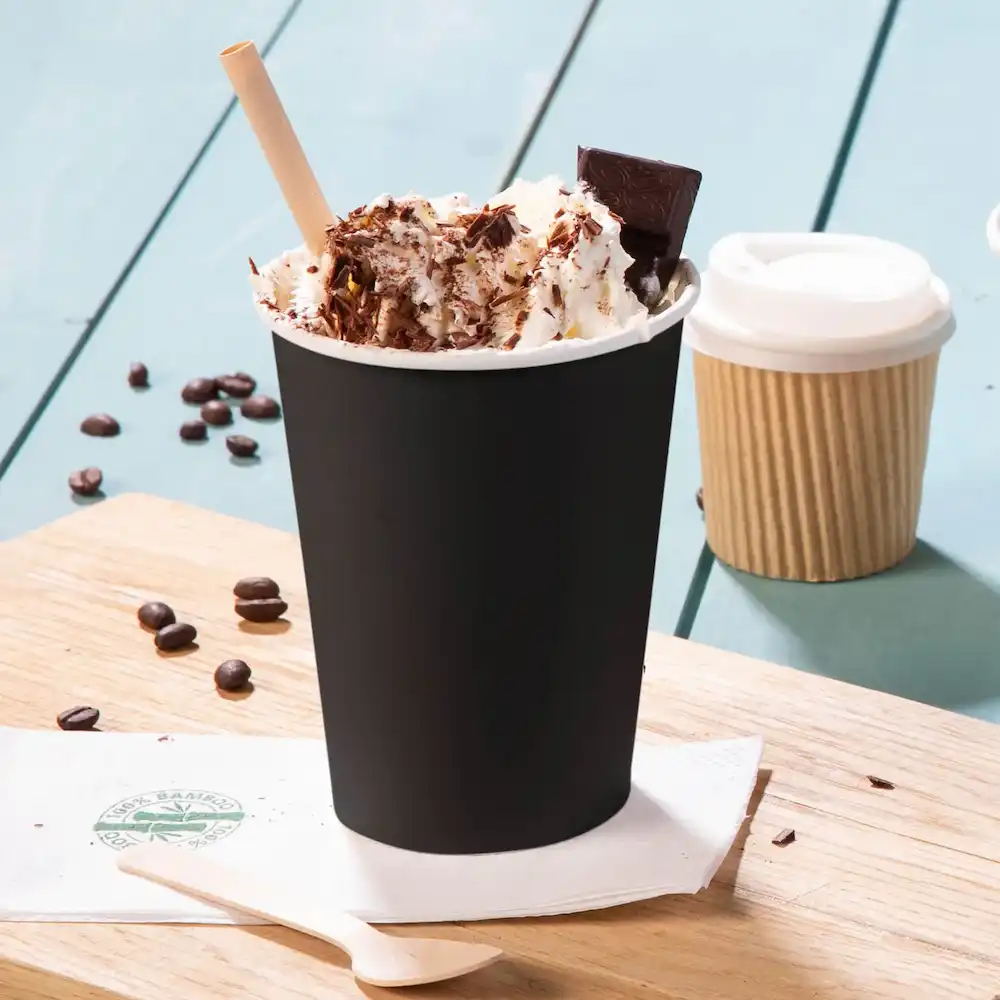 Black paper cup filled with whipped cream, chocolate shavings, and a chocolate square, with coffee beans scattered nearby.