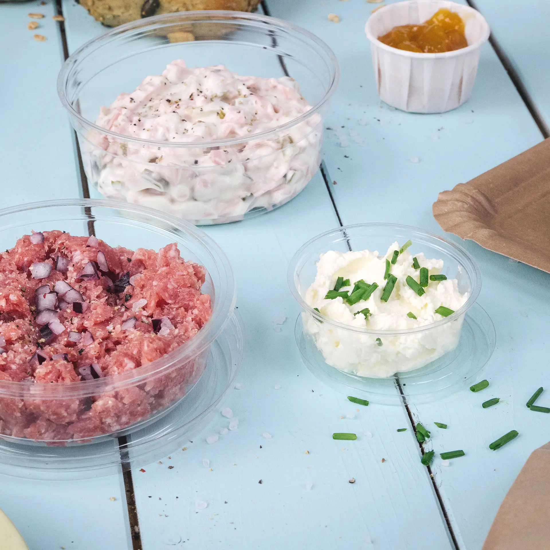 Three transparent portion cups containing different food items on a light blue wooden surface. The cups include minced meat, white dip with chives, and a sauce in a small cup.