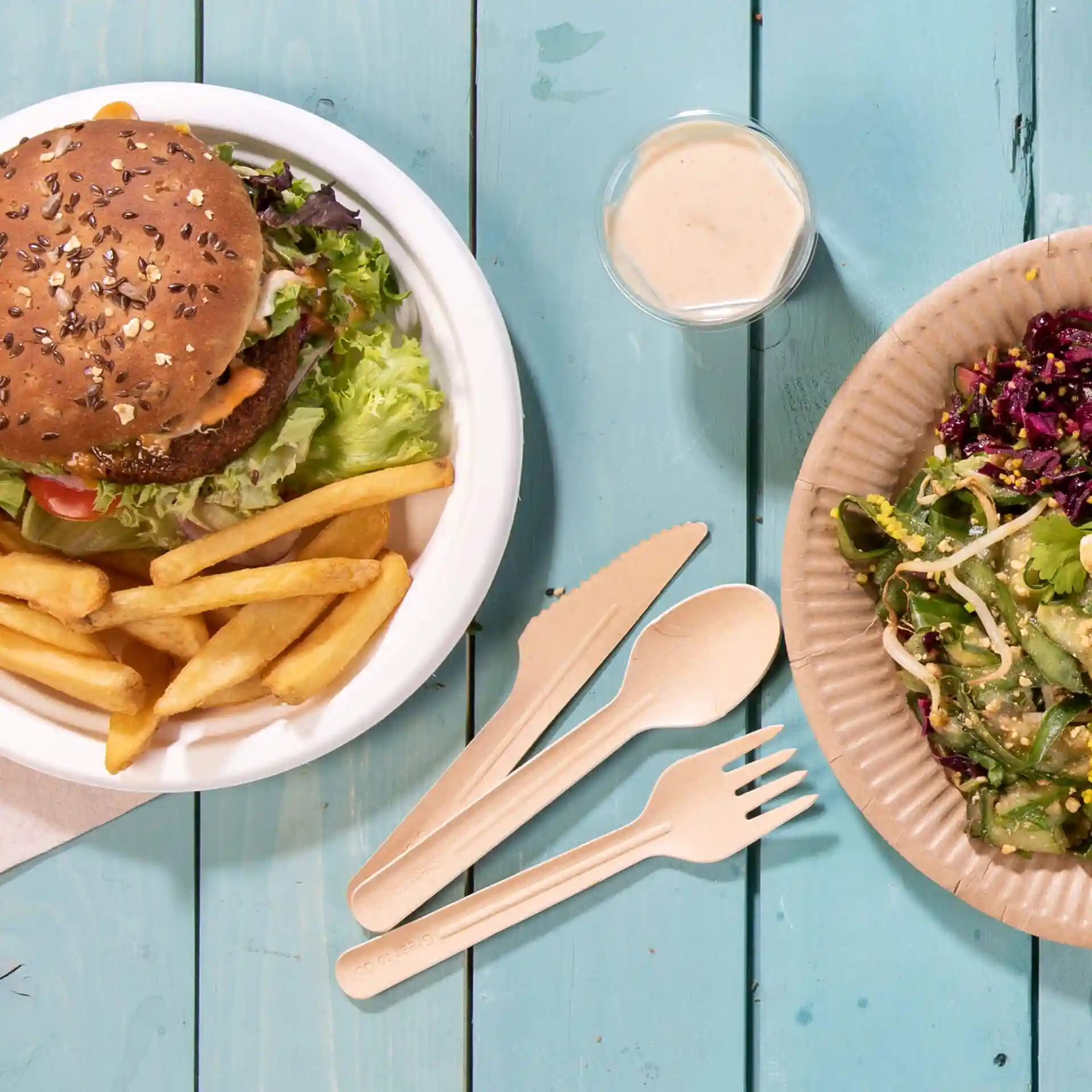 Image of a burger with lettuce and fries in a white bowl, alongside a salad in a brown paper plate and a small cup of dressing.