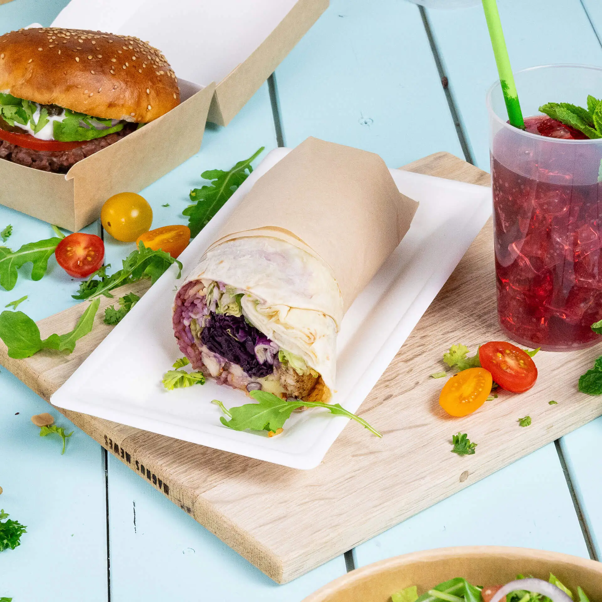 A rectangular sugarcane plate holds a wrapped food item with lettuce, and a beverage sits beside it on a wooden surface.