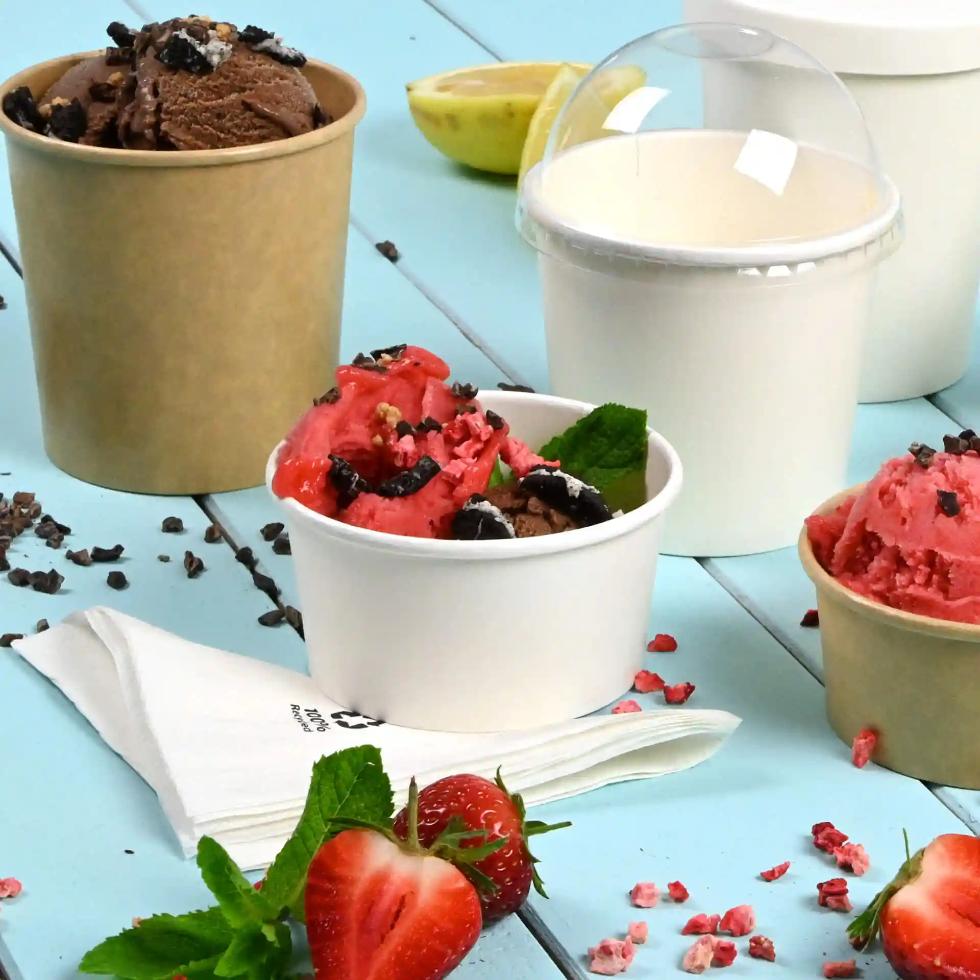Image of multiple paper ice cream cups in white and brown, filled with various ice cream flavors, with strawberries and mint leaves.
