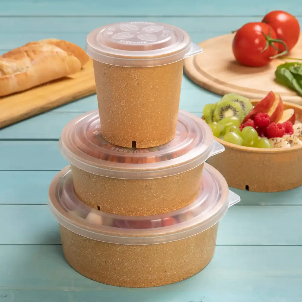 Stack of three brown containers with clear lids on a blue surface, with bread, tomatoes, and fruit salad nearby.