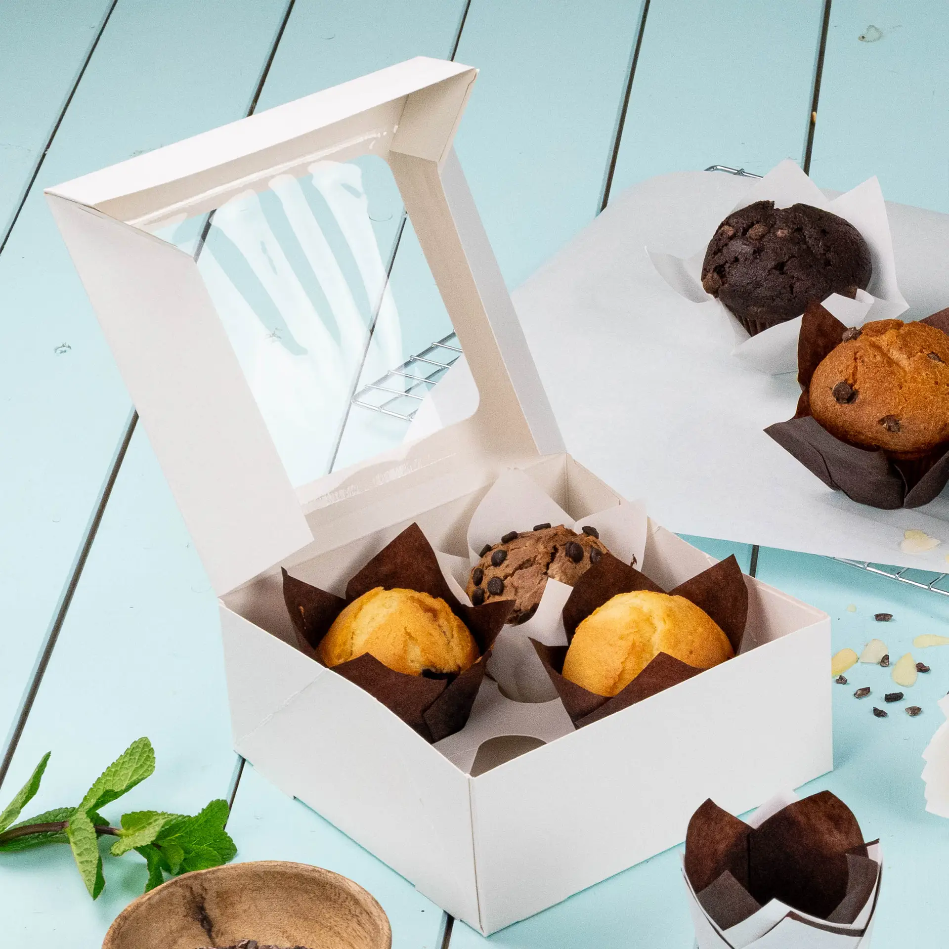 White 4-piece cupcake box with a cellulose window, containing muffins and a chocolate chip muffin, on a light blue surface.
